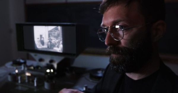 A man with beard and glasses sits in front of a editing machine that is playing a black and white movie.