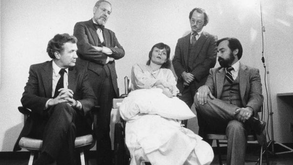 Black and white photo. A woman sits in a wheelchair surrounded by four men in suits.