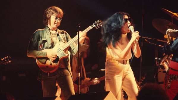 Yoko One on stage in a white top and pants singing into a microphone while John Lennon plays guitar behind her.