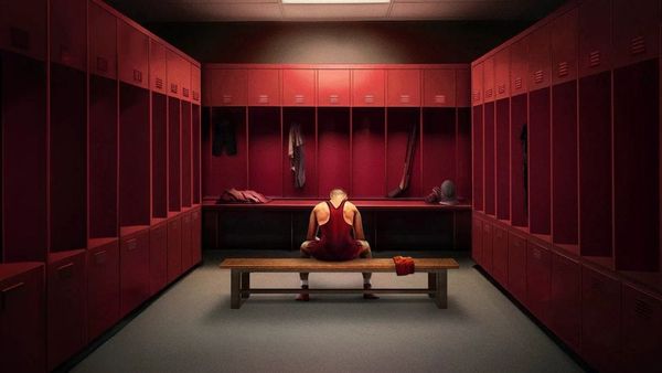 A young man in a wrestling uniform sits in a stylised locker room where everything is the colour red.