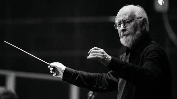 A black and white image of an ageing man in a dark outfit conducts an orchestra.