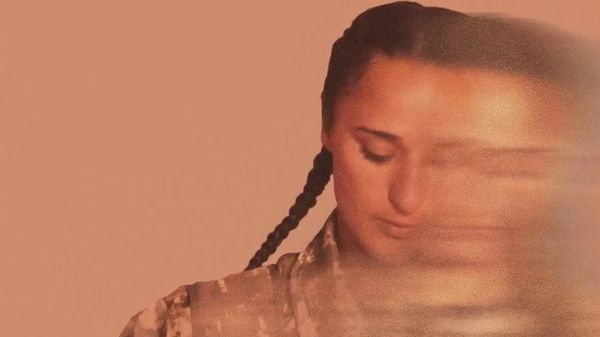 A woman in army fatigues and plaited hair looks contemplatively.