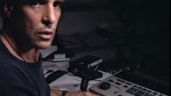 A man looks at the camera, turning from his desk covered in technical equipment.