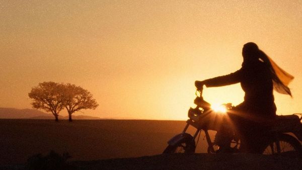 A woman rides a motorcycle against a sunset.
