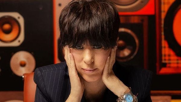 Diane Warren sits in front of a wall of speakers and stares directly into the camera with her hands on her face.