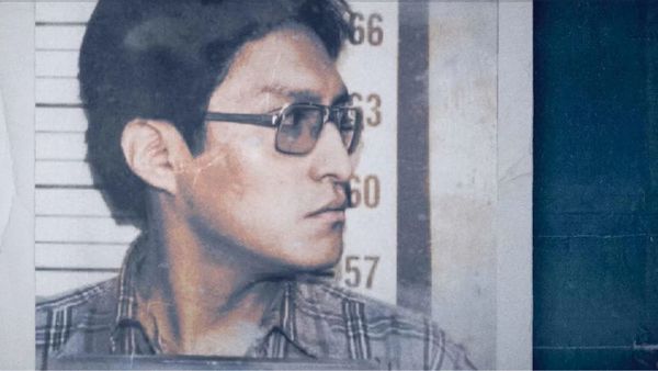 An Apache man in glasses and a plaid shirt stands in front of a police height chart looking to his side.