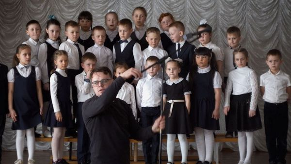 A teacher in black adjusts a microphone in front of a classroom choir of young children.