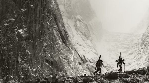 Two people hike with skis on their face as massive mountain faces appear behind them.