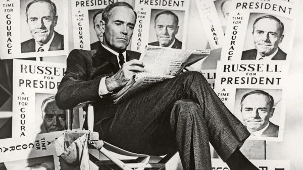 Henry Fonda in a suit and tie sits cross-legged reading a newspaper surrounded by signs that read Russell for President, a time for courage.