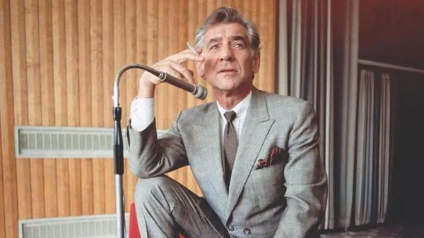 Leonard Bernstein in a great suit with brown tie, smoking a cigarette in front of a microphone in a recording studio.