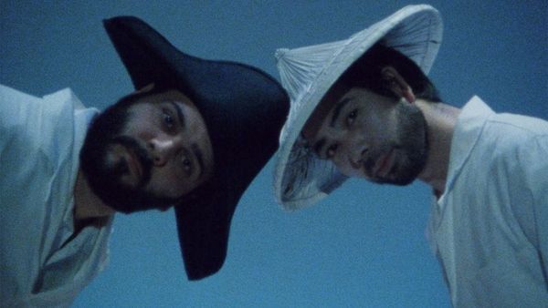 Two men, one with a beard in a black pirate-like hat and the other, an asian man in a white hat, turn the camera.