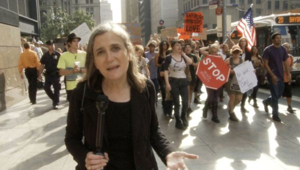Amy Goodman, dressed all in black and holding a microphone, stands in front of a protest.