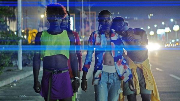 Four black dancers wearing brightly-coloured clothes on a street as car headlights shine around them.