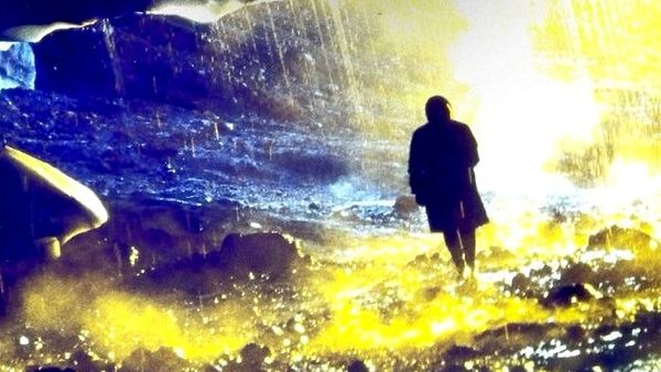 A silhouetted figure walks through a cave dripping with water coloured by yellows and blues.