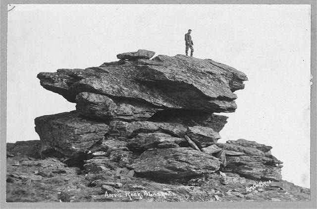 Old photograph of a man standing alone on top of a large rock formation.