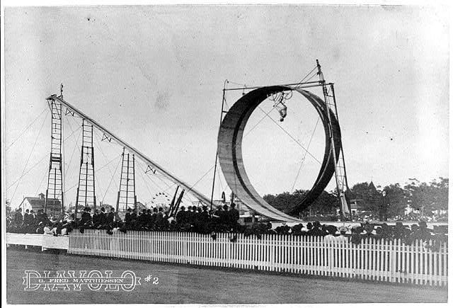 In an old photo, a man shown upside-down on a bicycle as he rides through a loop-the-loop stunt course.