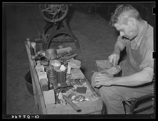 Old black and white photo of man at a workbench repairing a shoe.
