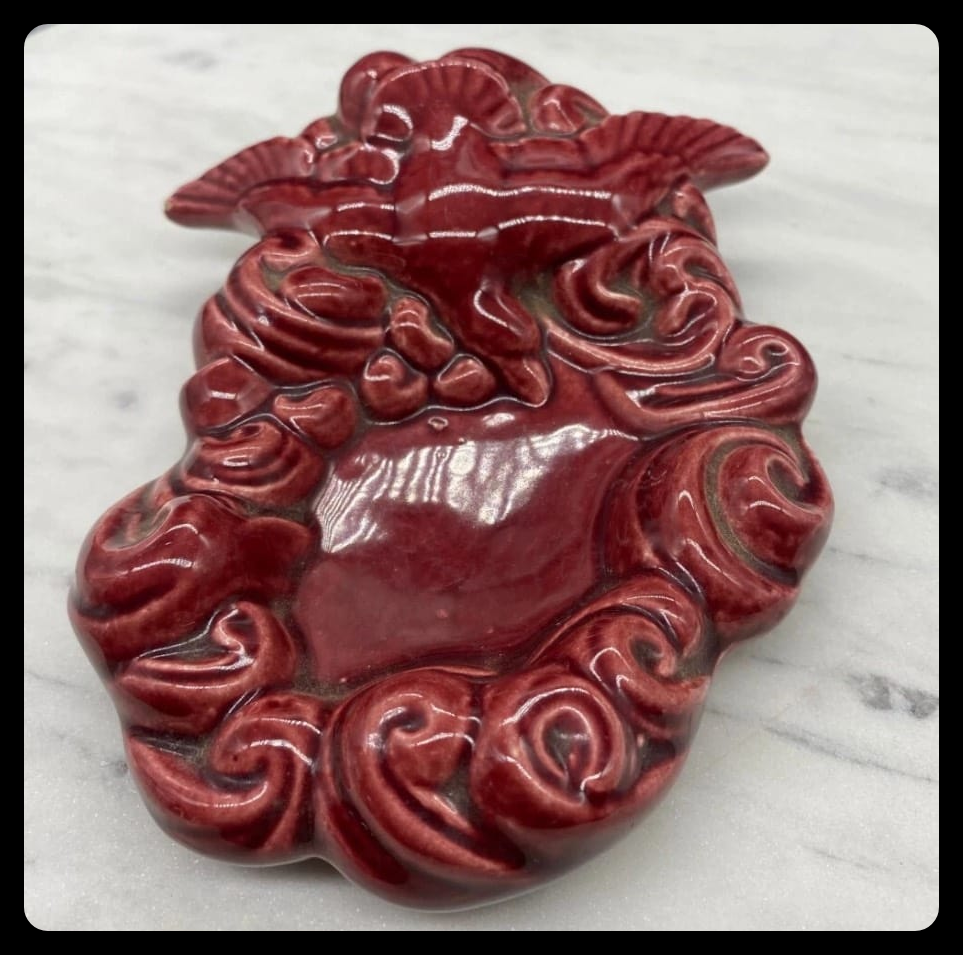 An ugly, old-fashioned mid century pottery trinket tray. A flying bird surrounded by swirling clould-like shapes. It's painted a lustrous red. 