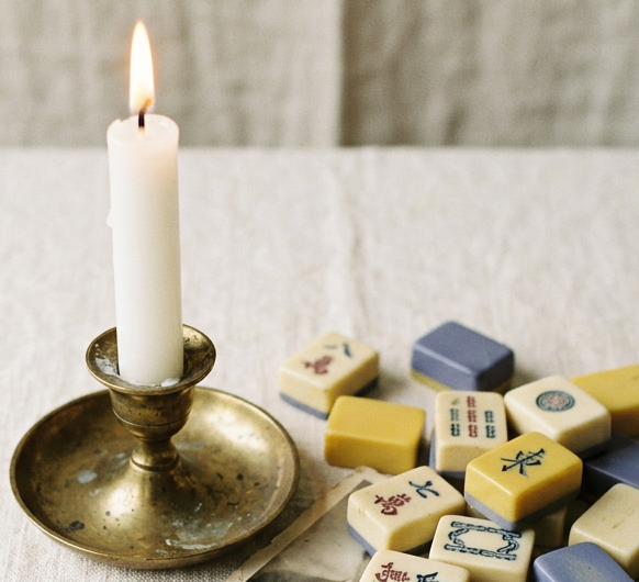 A lit white taper candle in an antique brass holder beside vintage mahjong tiles on cream linen.