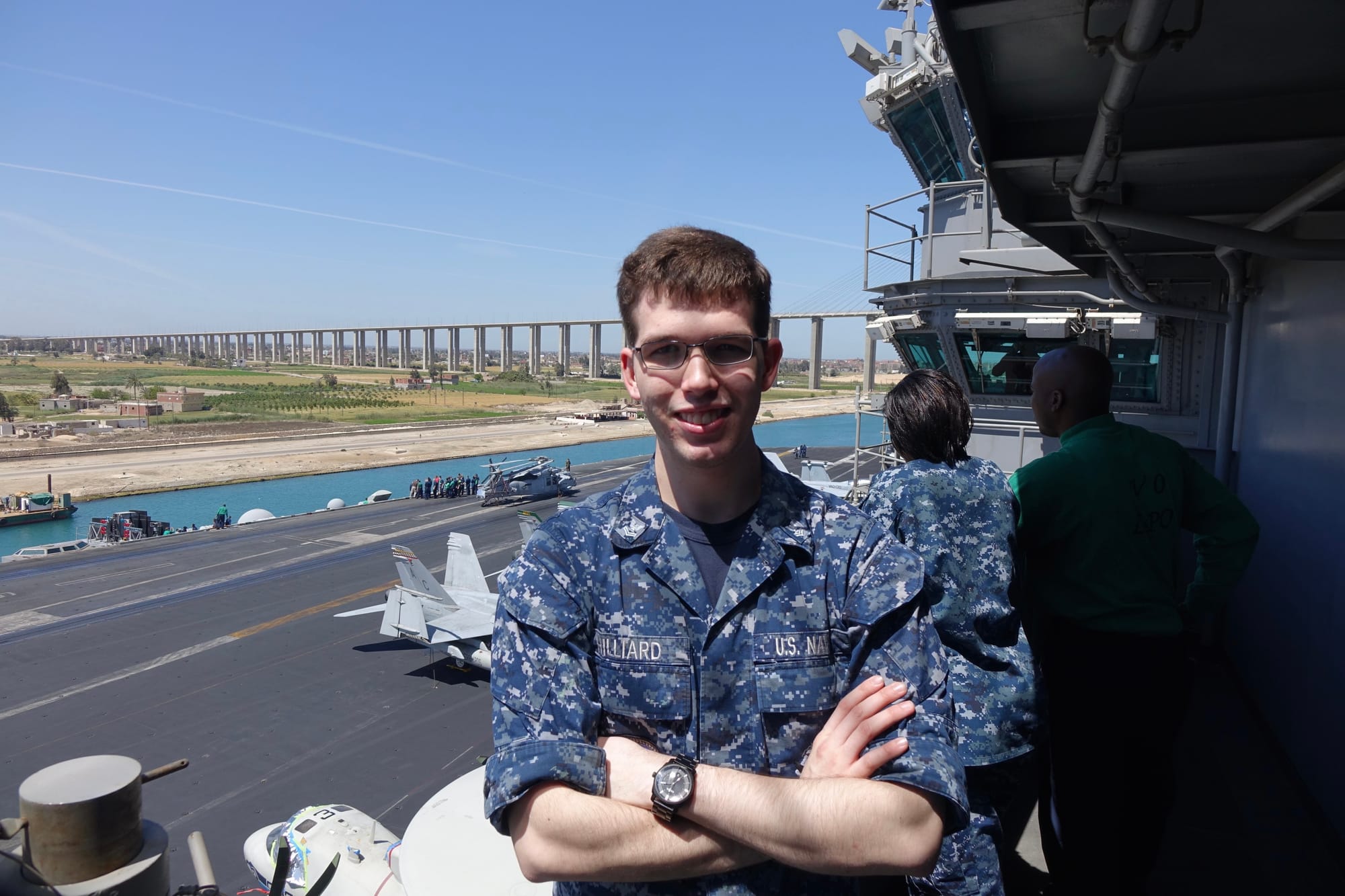 Wesley Hilliard in a blue camouflage Navy uniform standing on an aircraft carrier with a bridge in the background