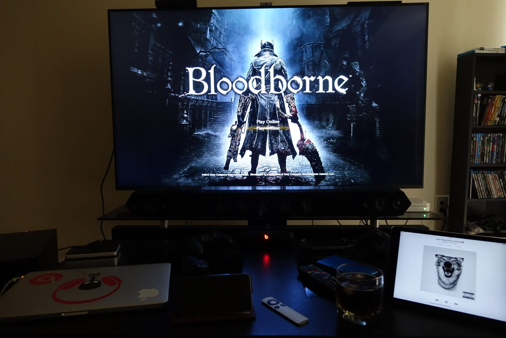 A photo of an apartment living room with a large TV showing the game "Bloodborne." A coffee table has an iPad, iPhone, MacBook Pro, and Apple TV remote