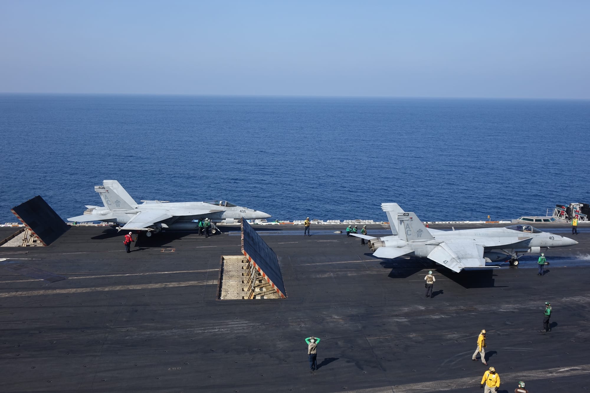 Two jets on an aircraft carrier being prepared for launch, visible airmen walking around the deck