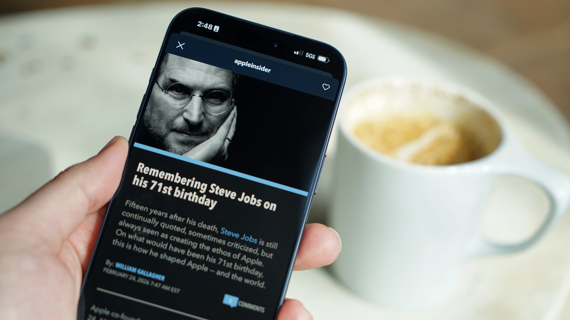 A post on the AppleInsider app about Steve Jobs shown on an iPhone held above a cup of coffee