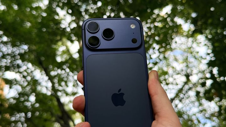 A deep blue iPhone 17 Pro Max held in a hand with trees in the background