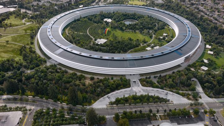 Apple Park, the giant ring campus in Cupertino, viewed from above and to the side, the rainbow stage visible in its center.