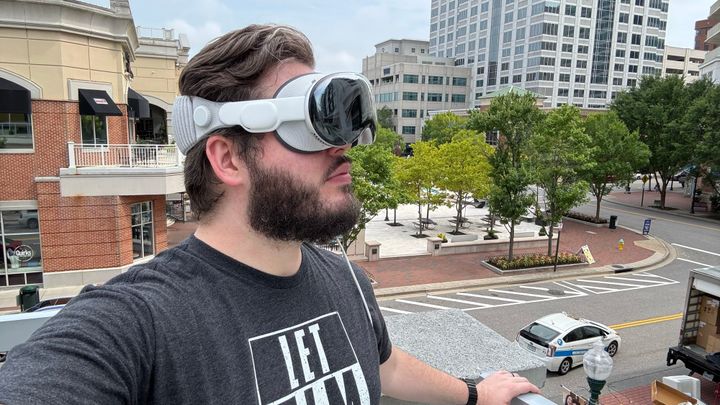 Wesley standing near Town Center in Virginia Beach wearing an Apple Vision Pro, trees in the background surrounded by buildings.