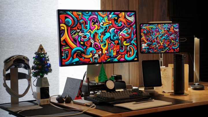 A desk with a Studio Display Monitor by an iPad Pro, iPad mini, Apple Vision Pro, keyboard, and various accessories in front of a window