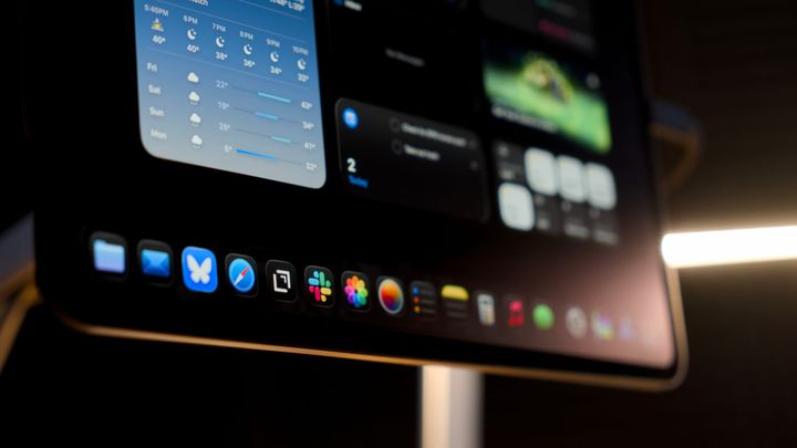 An iPad Pro on a magnetic stand shown with a desk lamp in the background, icons are visible in the dock and widgets on the Home Screen