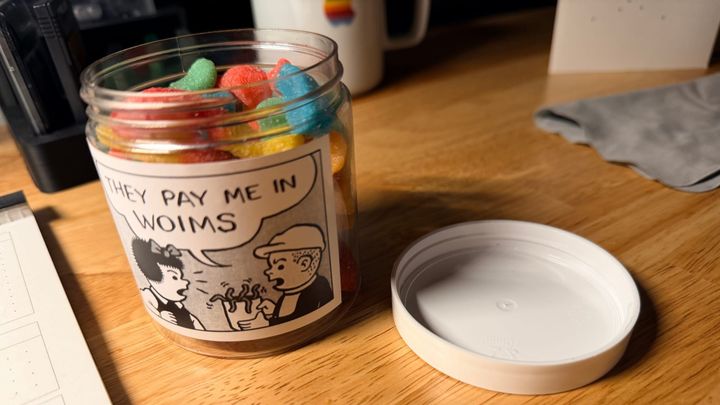 A plastic jar of gummy worms with a black and white comic of two characters, one holding a bucket of worms saying "they pay me in woims"