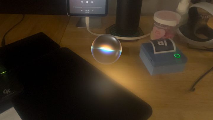 A view of a desk seen through Apple Vision Pro with a virtual sphere representing Siri shown in the center