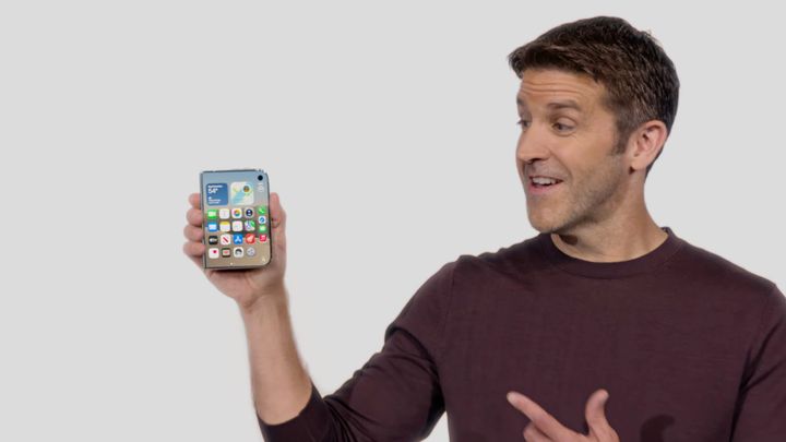 John Ternus in his maroon long sleeve shirt holding the closed iPhone Duo while pointing at it