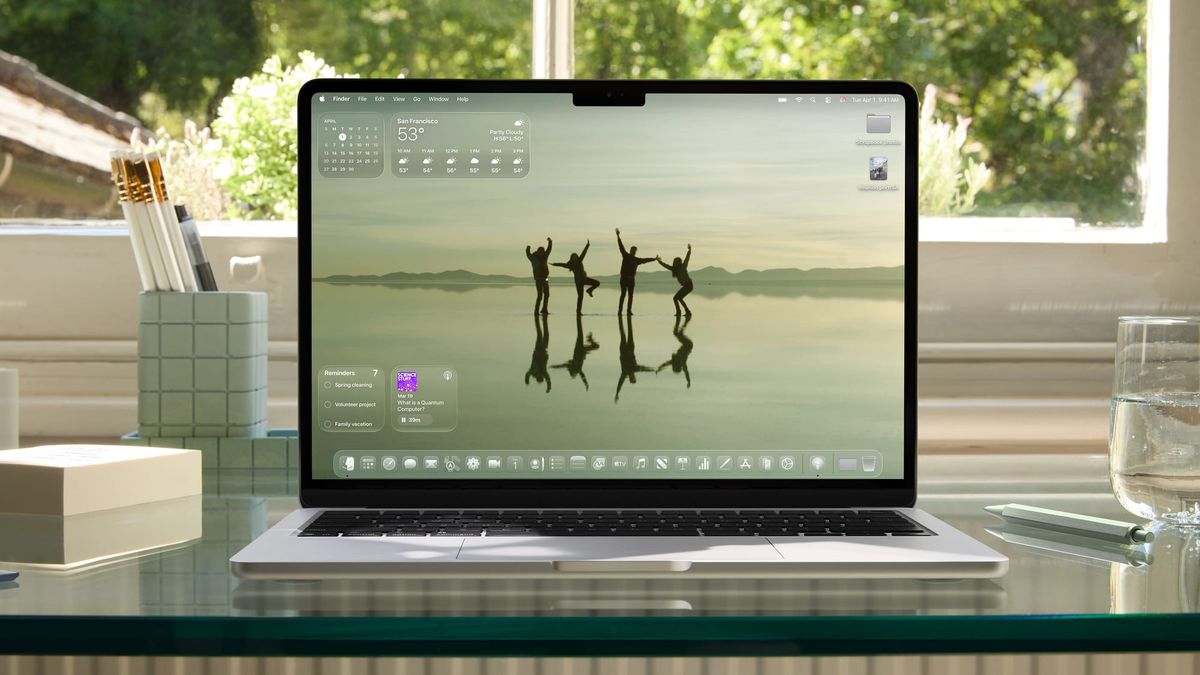 A MacBook Air on a glass desk with a window and trees in the background