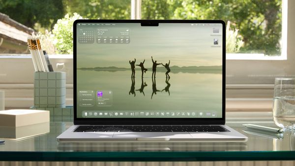 A MacBook Air on a glass desk with a window and trees in the background
