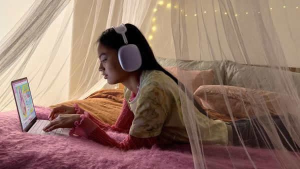 A young child with MacBook Neo in pink and AirPods Max (which cost almost as much as the laptop) in purple on a pretty fuzzy blanket in their bed