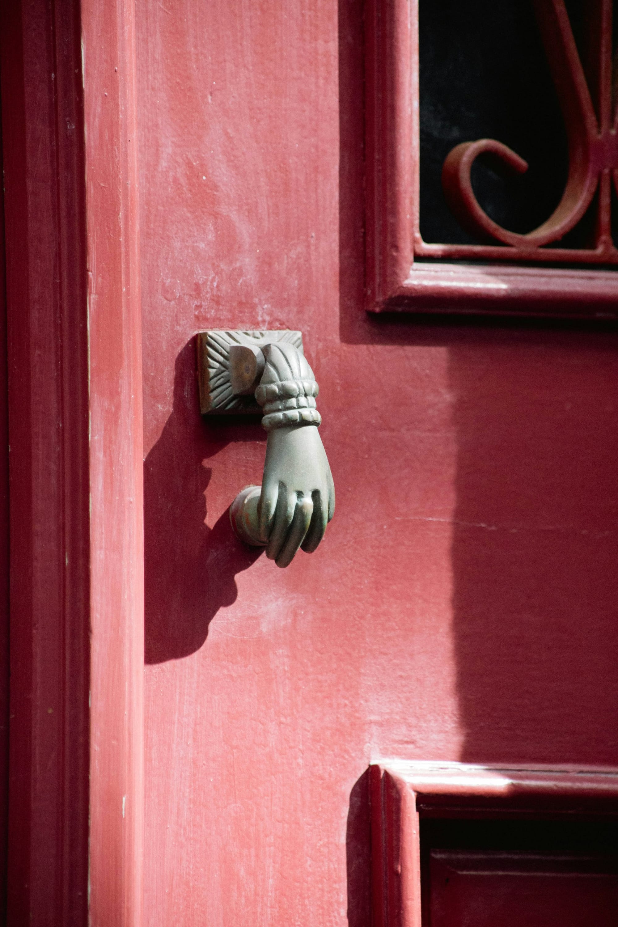 A vintage metal door knocker shaped like a hand holding a ball on a sunlit red painted wooden door.