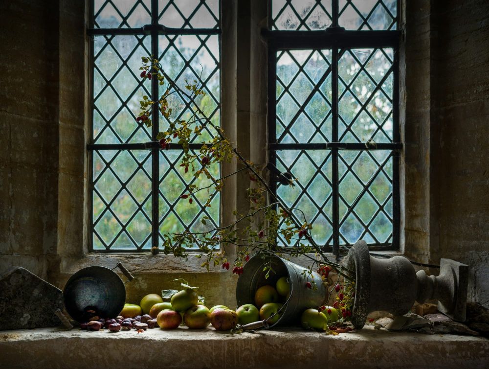 Apples, chestnuts and a tipped pail spill across a deep stone windowsill beneath leaded glass windows, with a sprig of rosehips and an old urn.