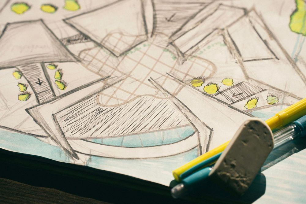 A hand-drawn pencil plan with hatched stairs and small painted shrubs lies on a desk beside a worn eraser and two pens.