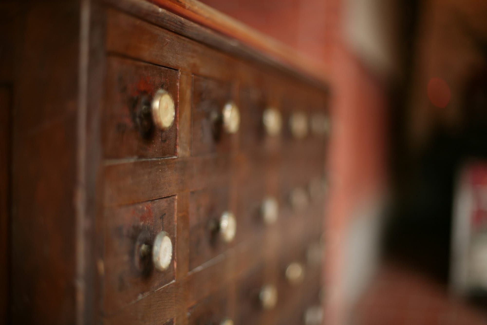 Rows of small drawers in an old worn wooden cabinet with pale round knobs and faint hand-marked labels, receding into soft focus.
