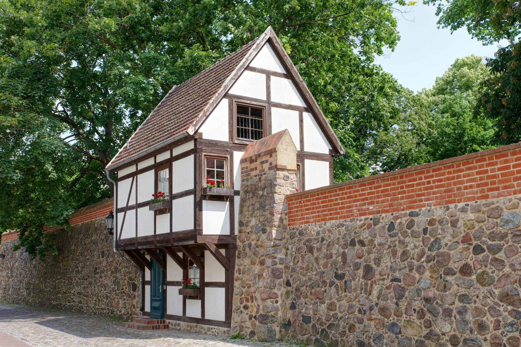 A narrow half-timbered house with red geraniums in its window boxes is built into an old fieldstone and brick town wall under tall trees.