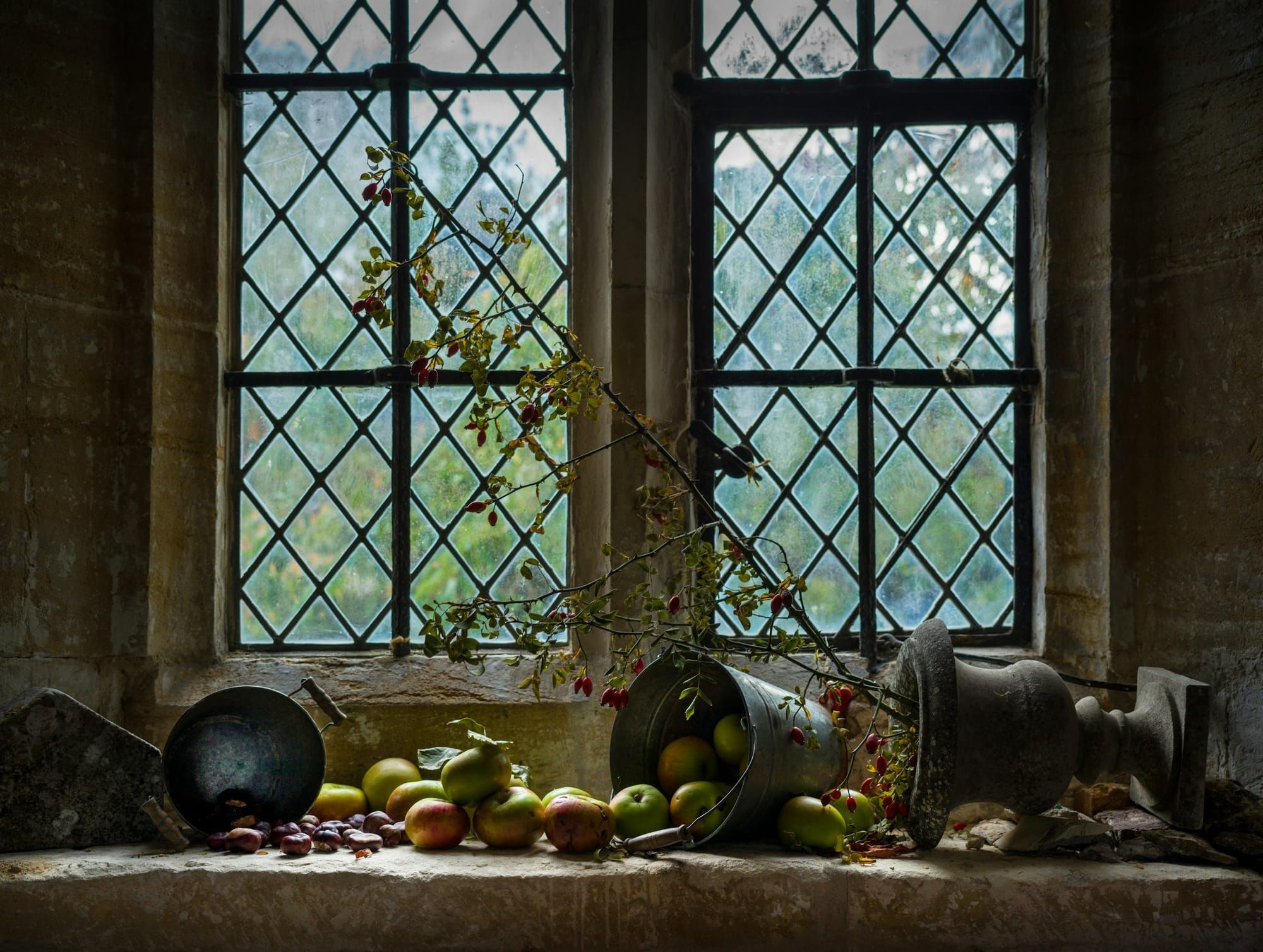 Apples, chestnuts and a tipped pail spill across a deep stone windowsill beneath leaded glass windows, with a sprig of rosehips and an old urn.
