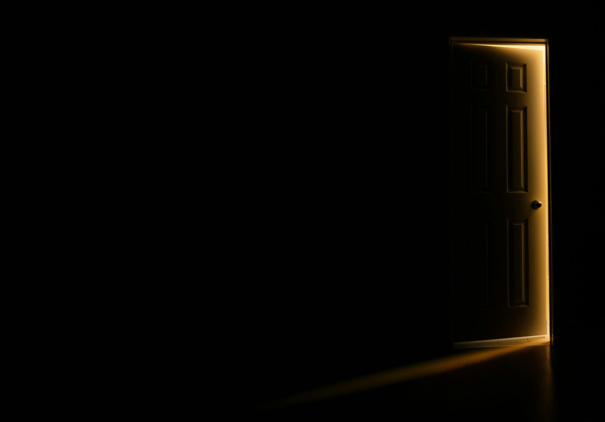 A panelled door stands slightly ajar in a pitch dark room, warm light spilling around its edges and across the floor.
