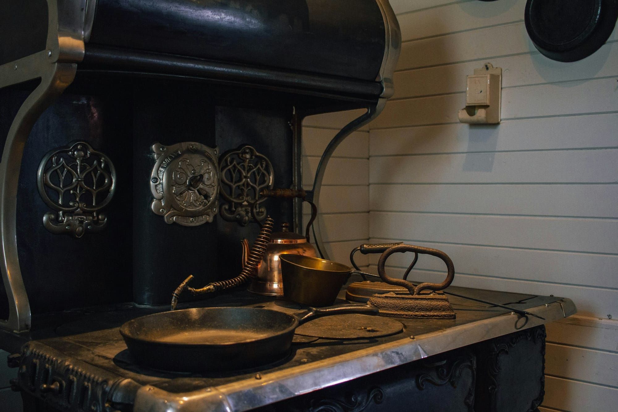 An antique cast iron kitchen range with ornate nickel trim holding a copper kettle and a brass cup and a skillet and old flat irons.