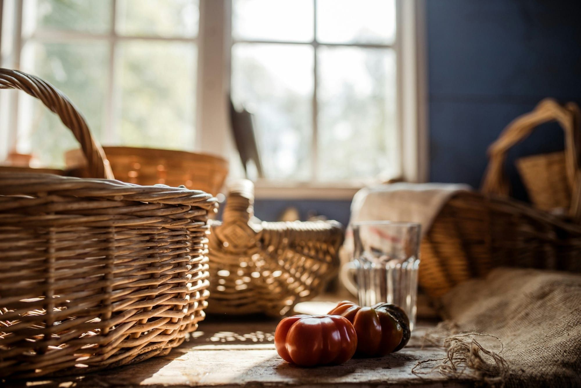 Ripe heirloom tomatoes sit on an old wooden table among wicker baskets and burlap in bright window light in a historic kitchen.