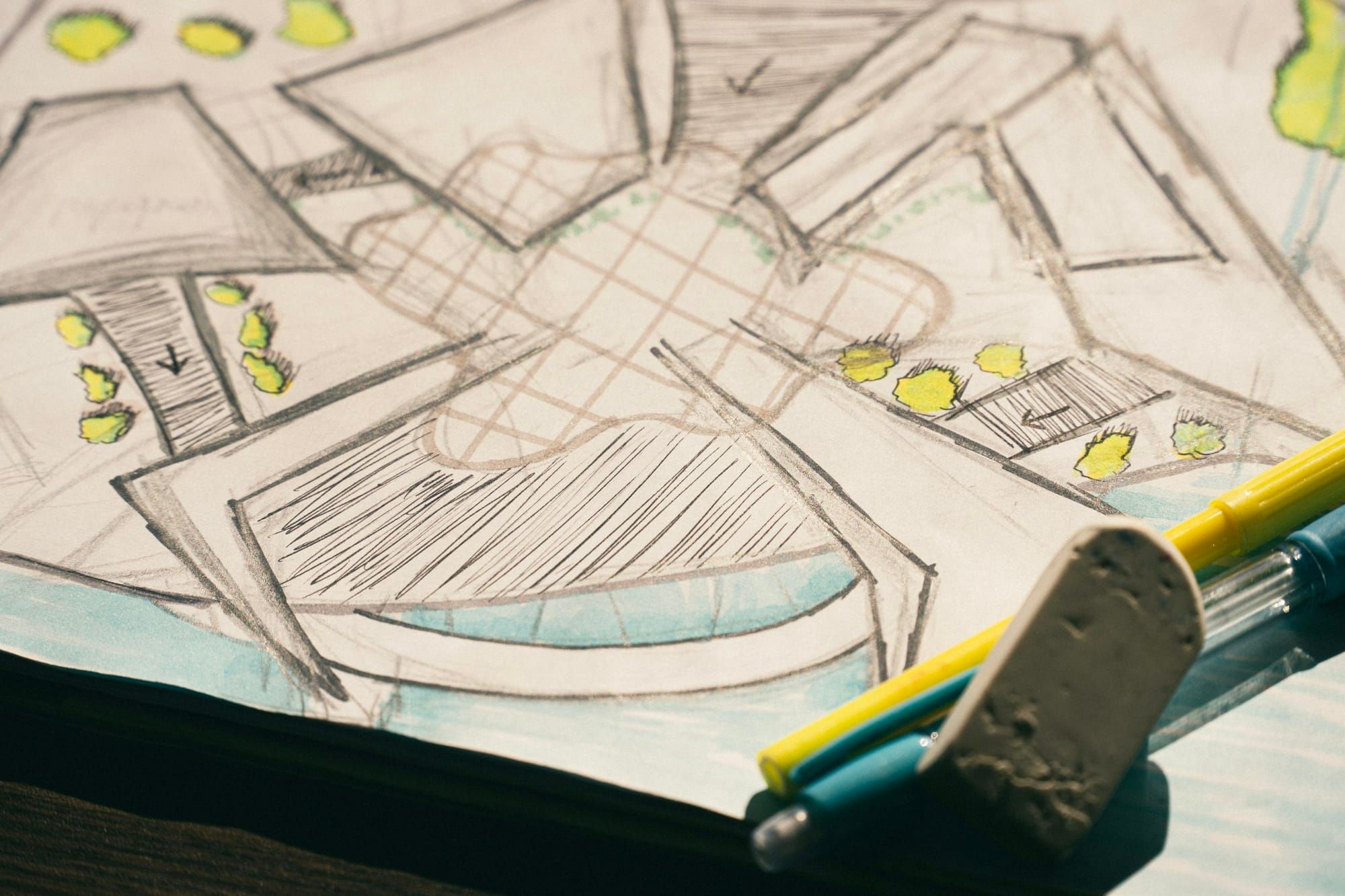 A hand-drawn pencil plan with hatched stairs and small painted shrubs lies on a desk beside a worn eraser and two pens.