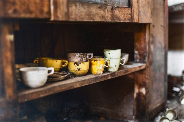 Chipped and mismatched old enamel cups in mustard yellow, cream and rust stand on a weathered wooden cupboard shelf.