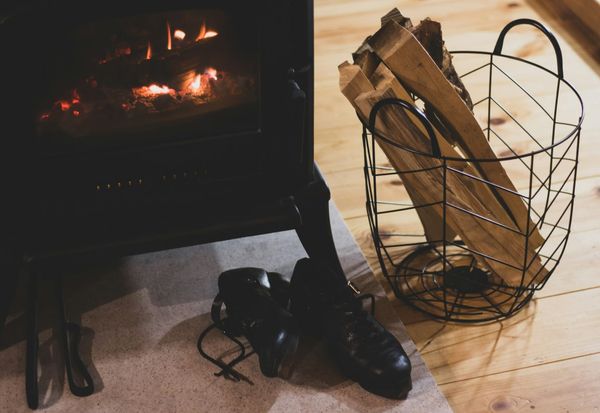 A pair of black lace-up boots rests on the hearth beside a lit wood stove and a wire basket of split logs.